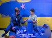 A woman and a child are sitting in a play area with large blue building blocks, smiling at each other. The background is a blue wall with starfish and cloud decorations.