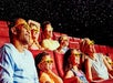 People wearing 3D glasses sit in a theater with red seats, watching a movie. They appear amused and engaged, as particles float in the air, suggesting an immersive experience.