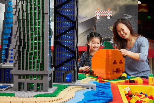 A woman and a young girl build a large orange LEGO pumpkin near a tall LEGO skyscraper at an indoor play area.