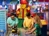 Two children and an adult assemble a LEGO set on a table in a brightly colored area, with a large LEGO figure in the background.