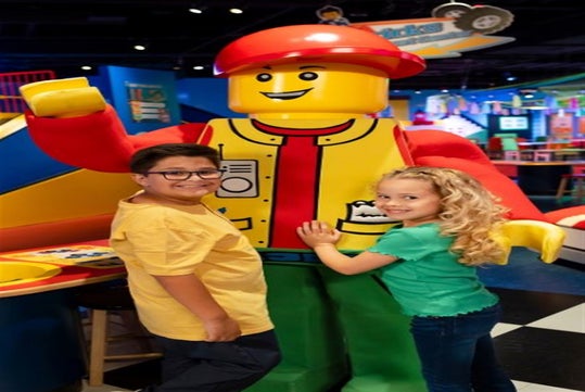 Two children smiling and standing beside a large LEGO figure indoors.