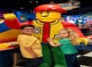 Two children stand next to and pose with a large LEGO character statue in a colorful indoor play area.