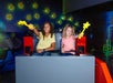 Two children sitting in a ride vehicle hold up yellow star-shaped wands, surrounded by a backdrop of glowing stars and space-themed decorations.