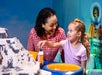 An adult and a child play together with LEGO bricks at an indoor activity table, with a large LEGO structure and colorful background visible.