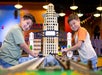 Two young boys smile while playing with a large LEGO city model featuring tall buildings and a bridge, inside a brightly lit indoor space.