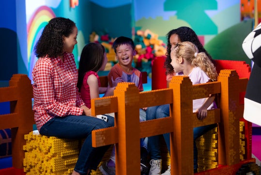 A group of children and an adult sit on colorful oversized toy bricks, smiling and talking in a brightly decorated play area.