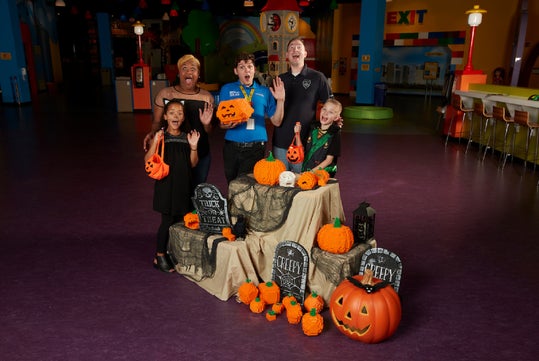 Five people pose and wave behind a Halloween display with pumpkins, gravestones, and decorations in a colorful indoor setting.