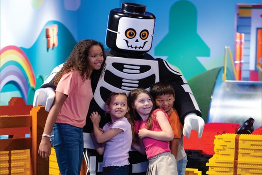 Four children smile and pose with a large skeleton-themed LEGO character in a colorful indoor play area decorated with rainbows and building blocks.