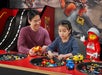 An adult and a child build with LEGO bricks at a play station, surrounded by various LEGO pieces and a LEGO figure in the background.