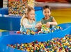 Two young girls are building with colorful LEGO bricks at a play area filled with plastic LEGO pieces.