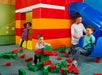 Three children play with large toy blocks and a slide inside a colorful indoor play area while an adult supervises.