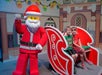 Two children sit in a red sleigh next to a person in a Lego Santa costume, indoors with festive decorations.