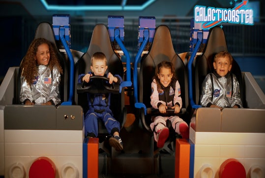 Four children in astronaut suits sit in a space-themed amusement ride labeled "Galacticoaster," smiling and holding on to the seat bars.