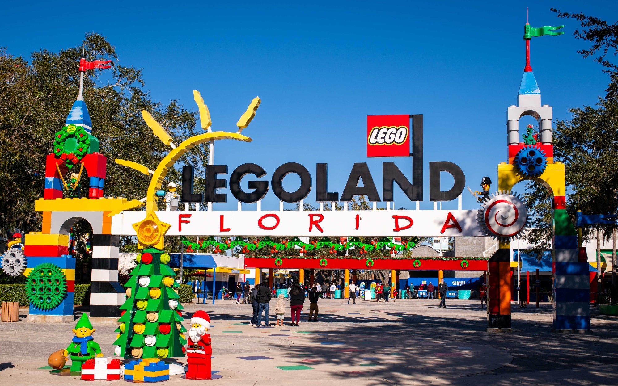 Entrance to LEGOLAND Florida featuring a large LEGO sign, colorful LEGO brick decorations, a Christmas tree, and visitors walking into the park.