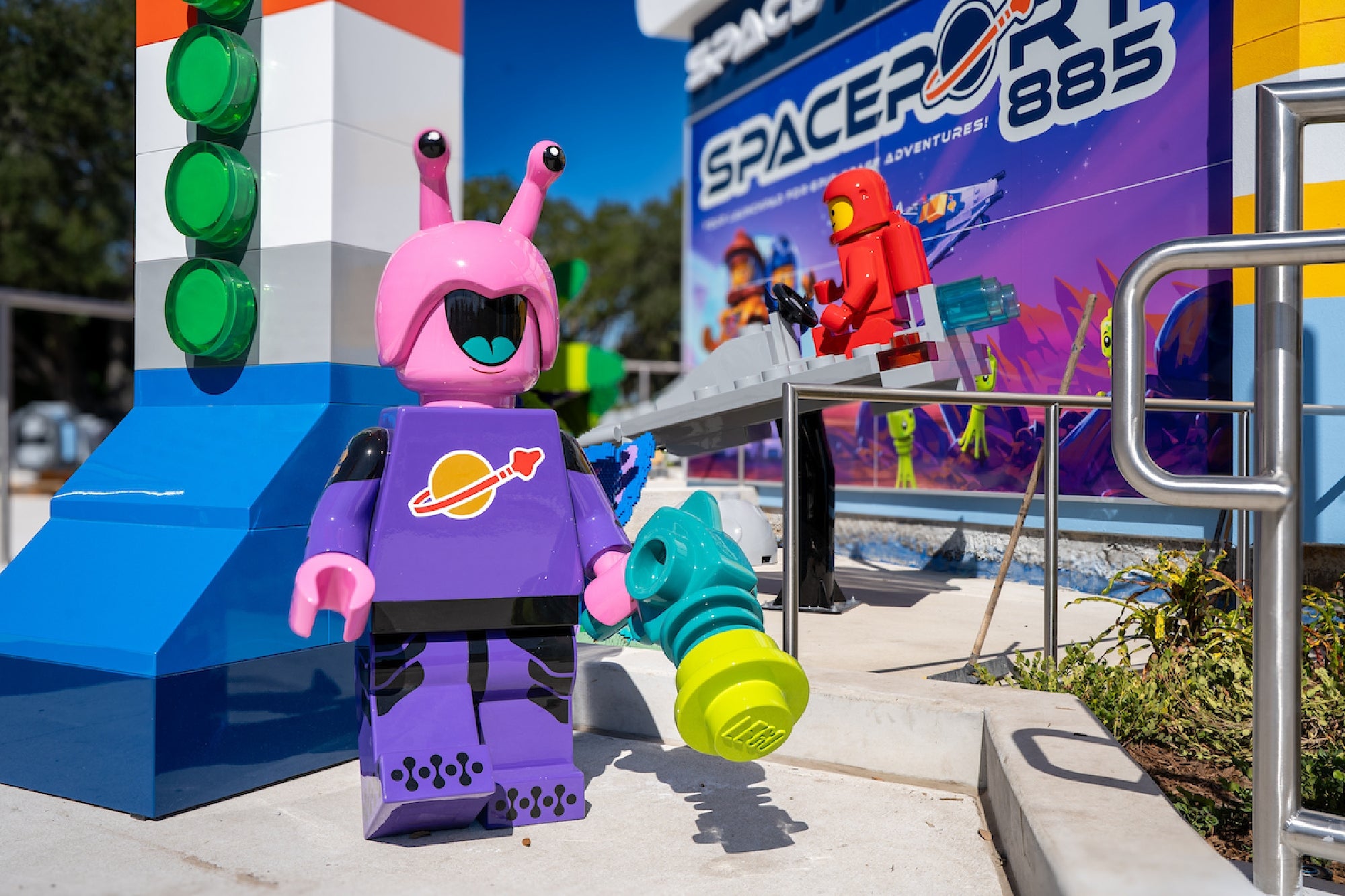 A pink and purple LEGO alien figure holding a toy blaster stands near a colorful "Spaceport 885" display, with a red LEGO astronaut figure in the background.