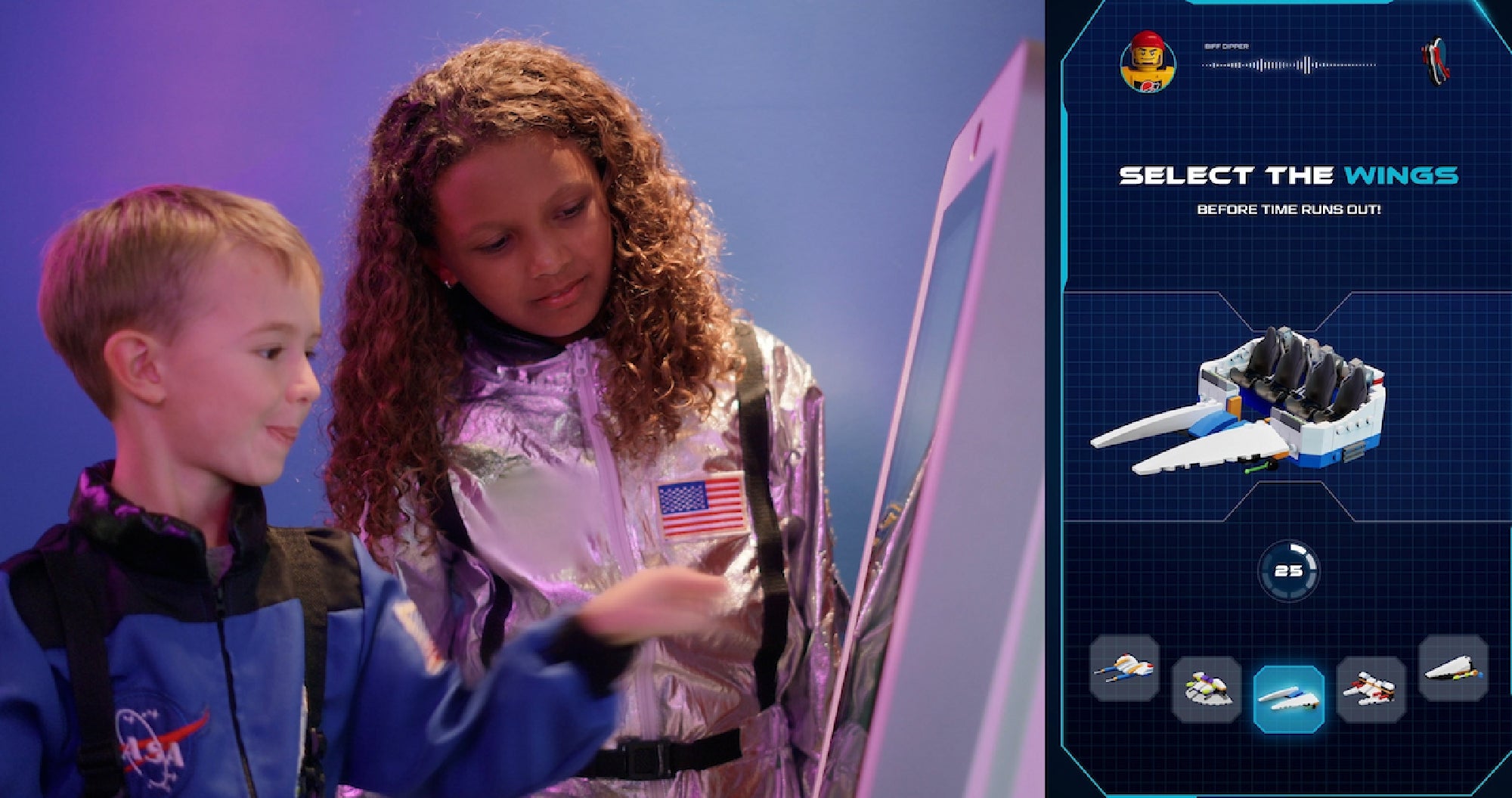Two children in NASA-themed outfits interact with a touchscreen, which displays instructions to select spaceship wings in a digital game.