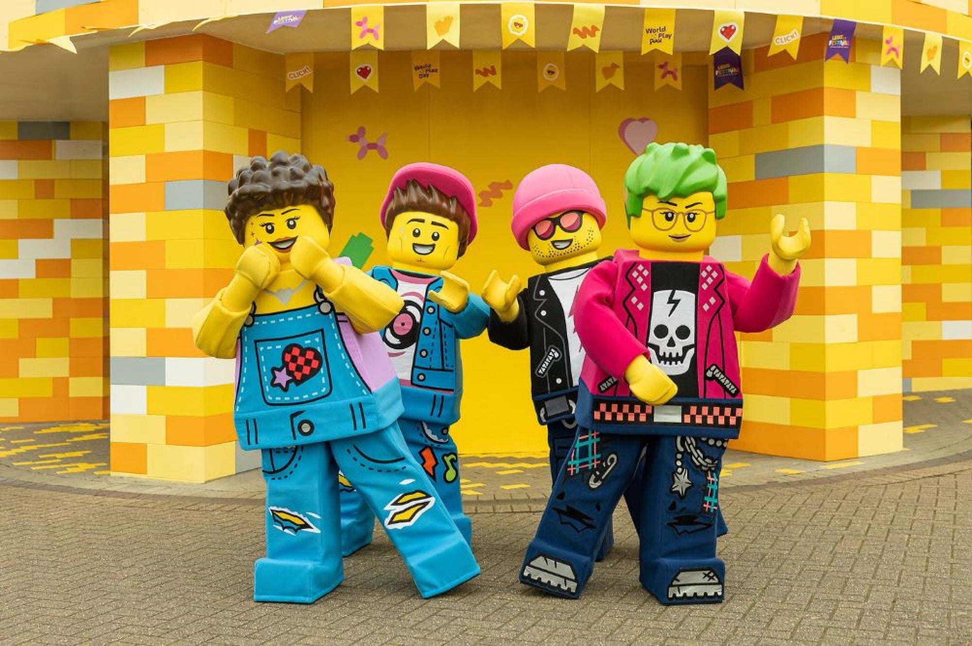 Four people in colorful LEGO character costumes pose in front of a yellow and orange building decorated with LEGO bricks and hanging flags.