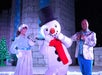 Two performers stand on stage with a person in a snowman costume. The background includes a snow-themed set with lights.