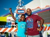 A man and a boy smile for the camera with arms raised in front of a colorful LEGOLAND attraction on a sunny day.