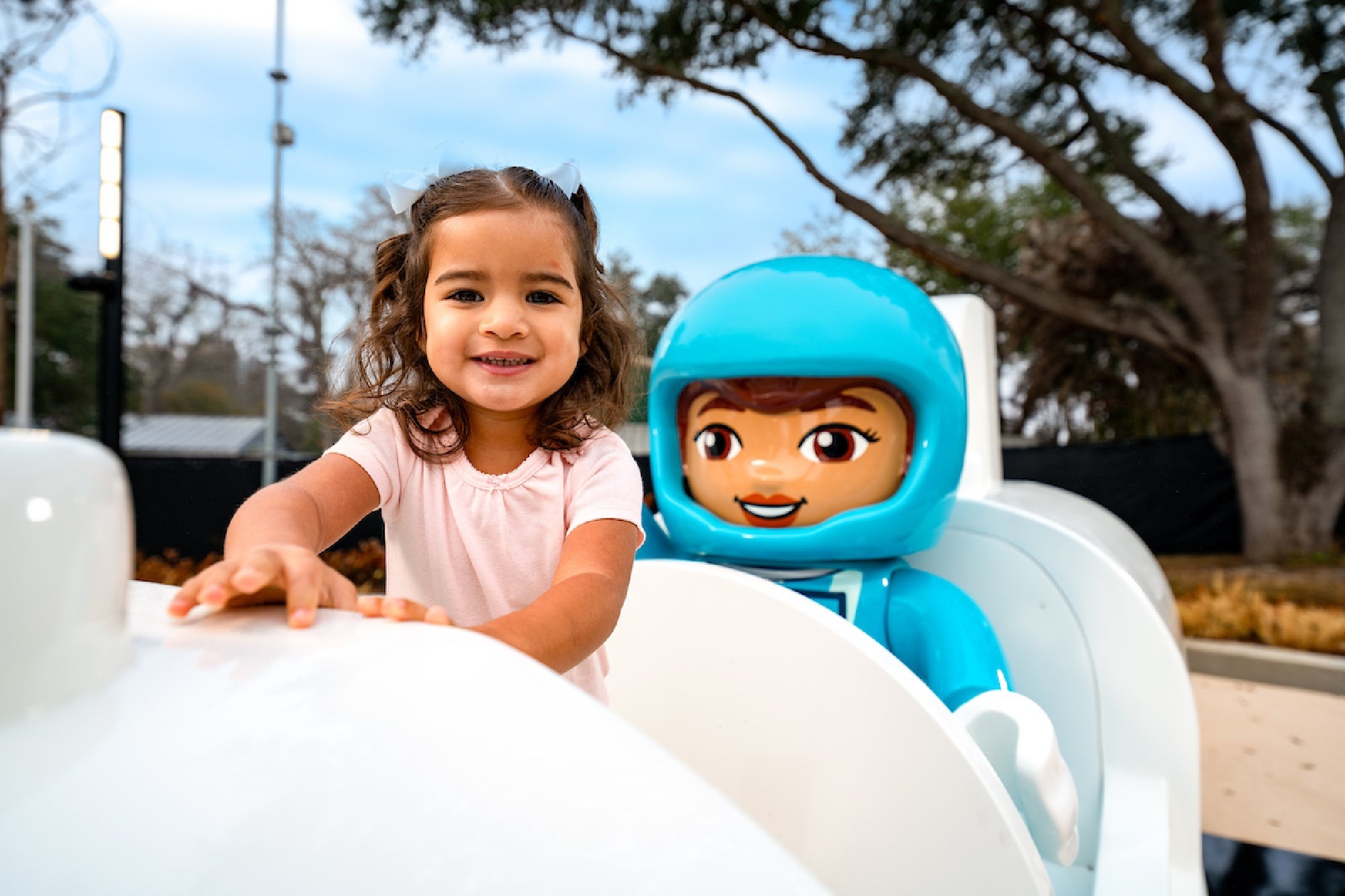 A young girl in a light pink shirt smiles while standing next to a large blue astronaut figure at an outdoor playground. Trees and sky are visible in the background.