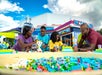 A family of four builds with large colorful toy bricks at an outdoor play area, with a building and blue sky in the background.
