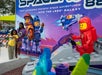 Two people pose for a photo in front of a colorful LEGO space-themed display with large LEGO astronaut figures and a spaceship mural in the background.