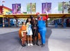 Four people in colorful costumes stand together posing for a selfie at an outdoor theme park with LEGO-themed decorations and banners in the background.
