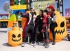 A group of people in Halloween costumes take a selfie next to large pumpkin decorations at an outdoor event called Monster Party.