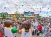 A crowd celebrates outdoors with confetti falling, surrounded by colorful buildings and two costumed characters resembling LEGO figures in the foreground.