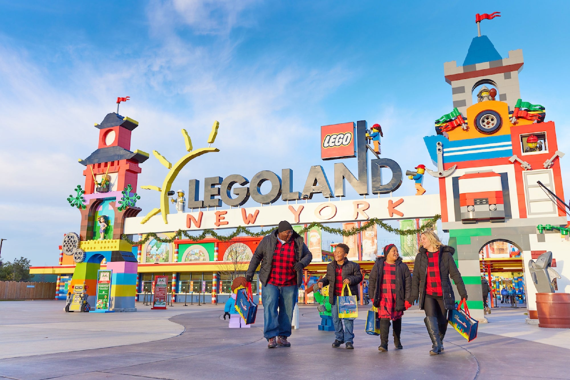 A family walks under the colorful entrance sign of Legoland New York, carrying shopping bags and wearing matching red and black plaid outfits.