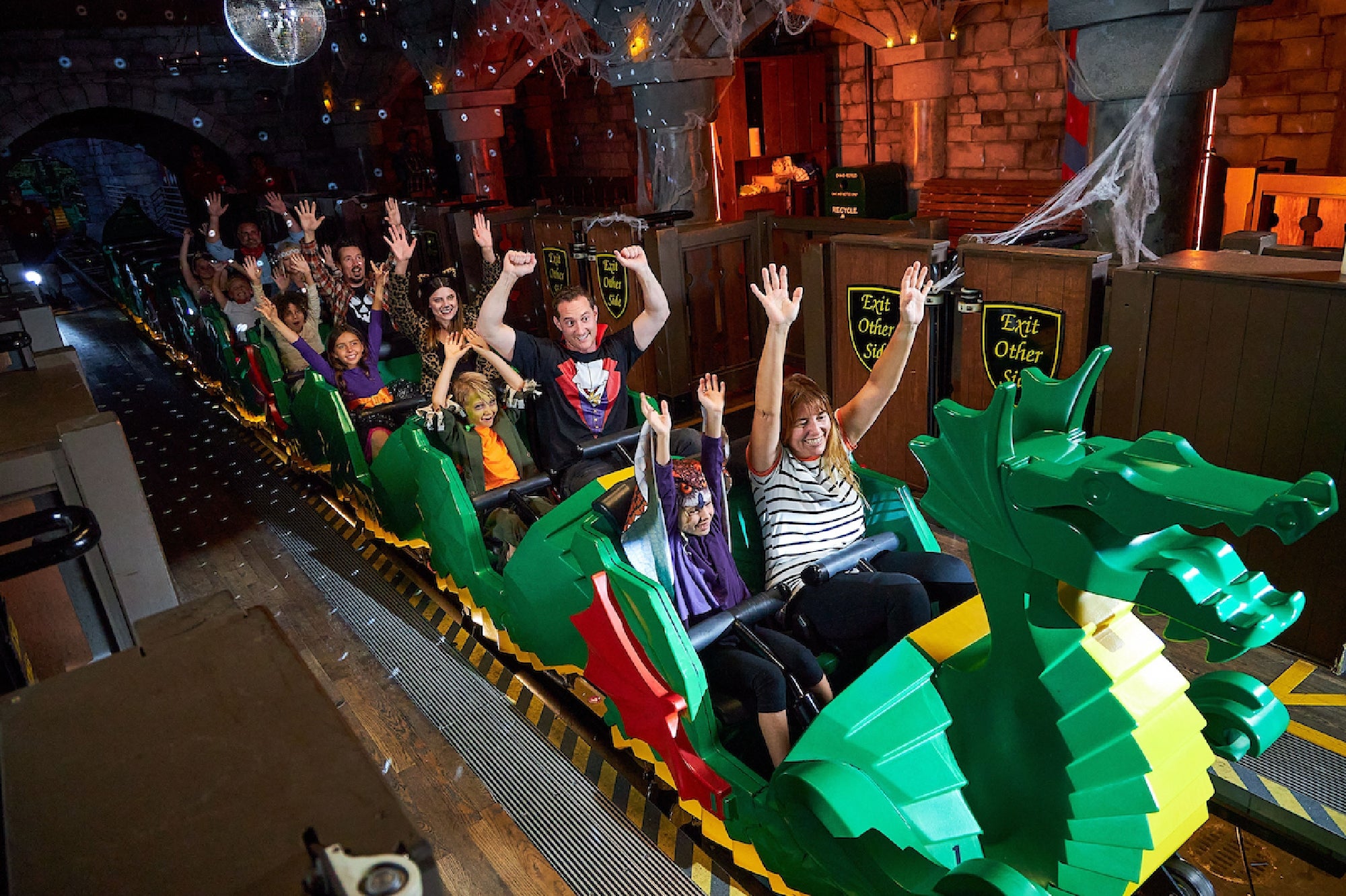 A group of people with raised hands ride a green dragon-themed roller coaster inside a dimly lit building with stone walls and cobweb decorations.