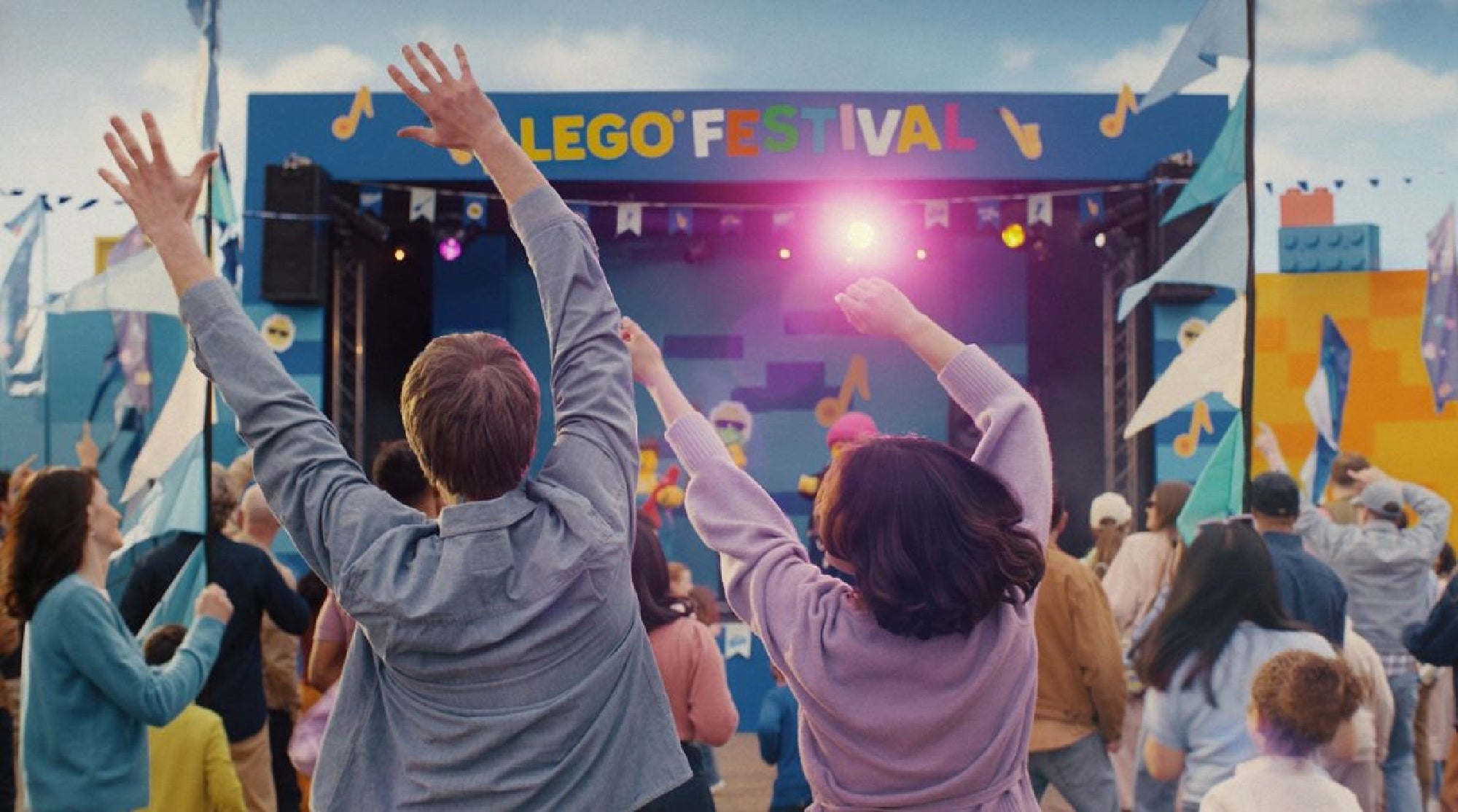 A crowd of people faces a colorful LEGO Festival stage, with many raising their arms and flags waving in the air under a partly cloudy sky.