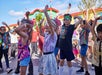 A group of children dance outdoors in colorful clothing, raising their arms, with a festive building and blue sky in the background.