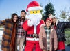 A family of four poses with a person in a Santa costume outdoors, near a decorated Christmas tree.
