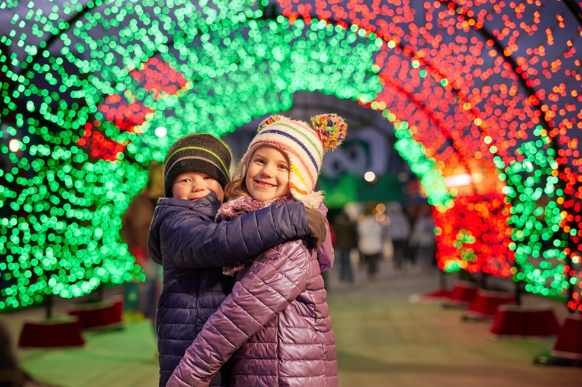 Two children wearing winter jackets and hats hug while standing under a tunnel of colorful holiday lights.
