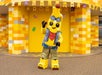 A person in a banana-themed costume with sunglasses, a vest, and shorts poses in front of a yellow and orange patterned building.
