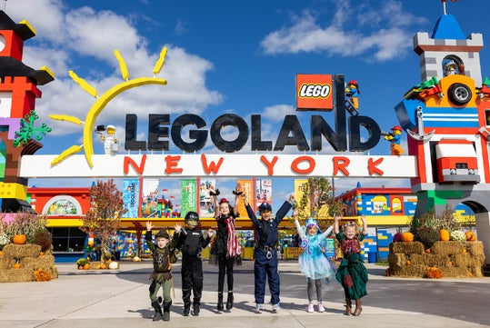Six children in costumes stand in front of the LEGOLAND New York entrance, with colorful Lego structures and clear blue sky in the background.