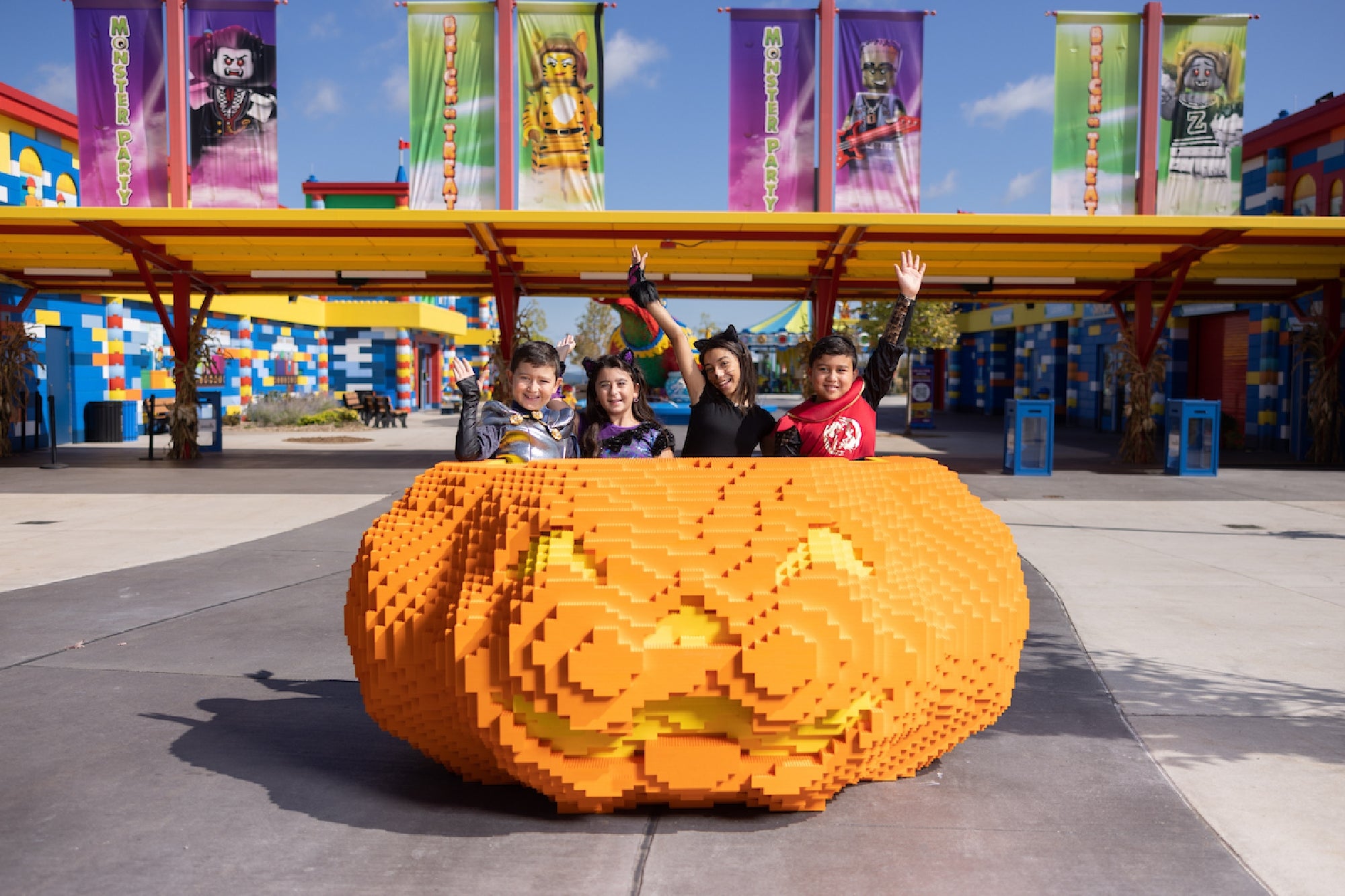 Four children sit and wave inside a large, orange, LEGO pumpkin sculpture at an outdoor amusement park decorated with colorful LEGO banners and buildings.