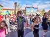 A group of children and adults dance outdoors at a colorful amusement park, with blue skies and a toy-like building in the background.