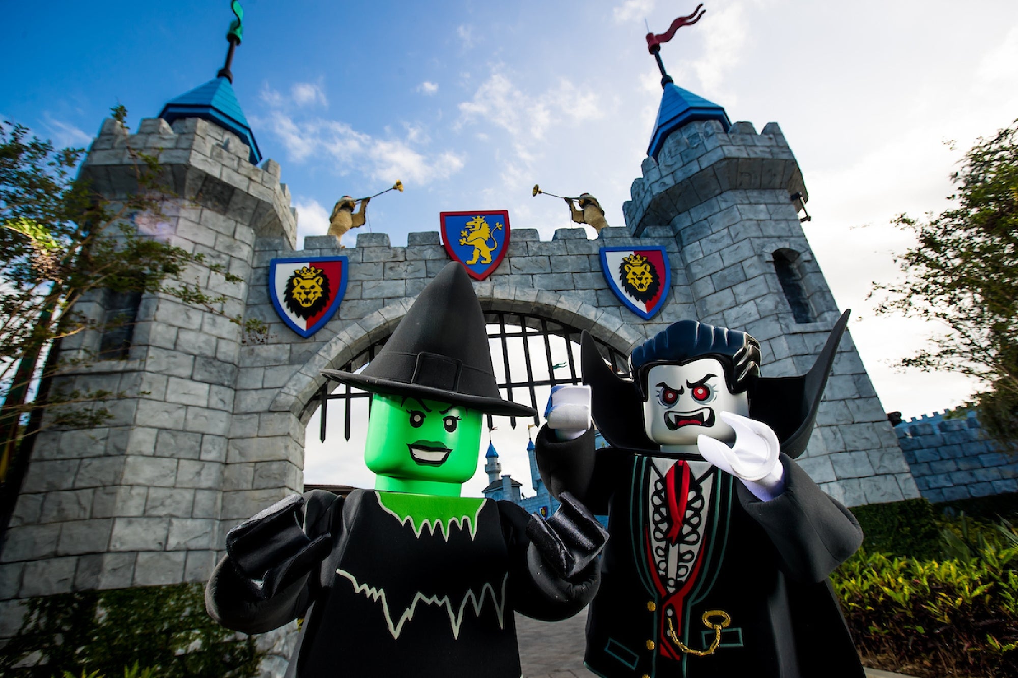 Two people in witch and vampire LEGO costumes pose in front of a castle entrance at a theme park, with blue towers and lion crests visible.