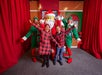 Two children and two people dressed as elves pose with a large Santa figure in a festive setting.