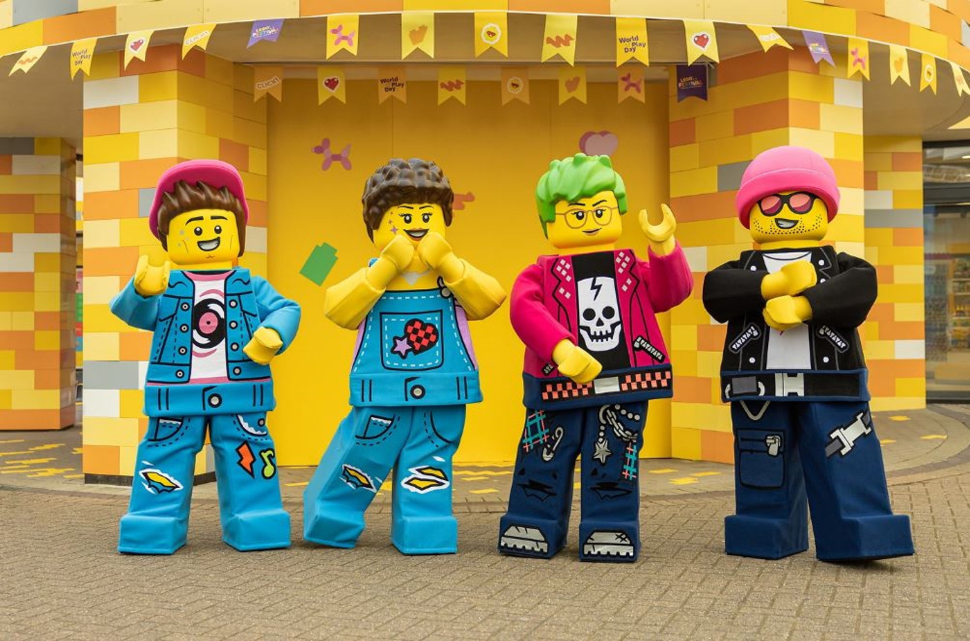 Four people in colorful LEGO minifigure costumes pose in front of a yellow brick-patterned building decorated with hanging flags.