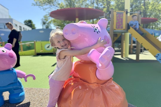 Young girl hugs a large Peppa Pig statue at a playground on a sunny day. Other children play in the background.
