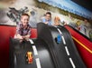 Two boys race toy cars down parallel, sloped tracks in an indoor play area with a backdrop of illustrated LEGO vehicles and a city skyline.