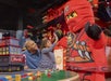 A child high-fives a person in a red LEGO ninja costume while an adult watches and smiles in a colorful LEGO-themed setting.