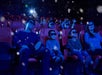 People wearing 3D glasses sit in a movie theater with red seats, watching a film as snow-like effects appear in the air around them.
