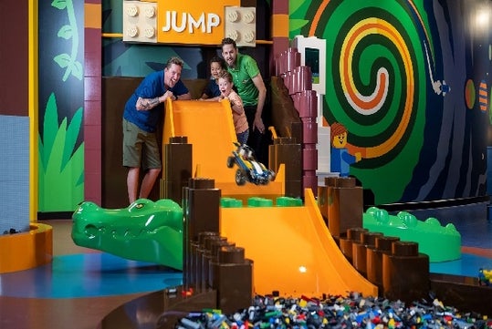 Two parents and two children at the top of an orange ramp with a giant LEGO alligator in the middle and a toy car in the air.