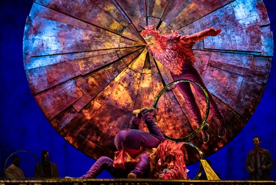 Performers in colorful costumes pose and tumble in front of a large, circular, textured backdrop on stage, with dramatic lighting and several people watching in the background.