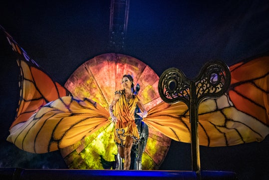 Performer in an orange butterfly costume poses on stage with large wings, standing in front of a circular, lit backdrop and stage prop.