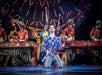 A performer kneels on stage juggling clubs, surrounded by musicians playing marimbas, with a large, colorful backdrop behind them.